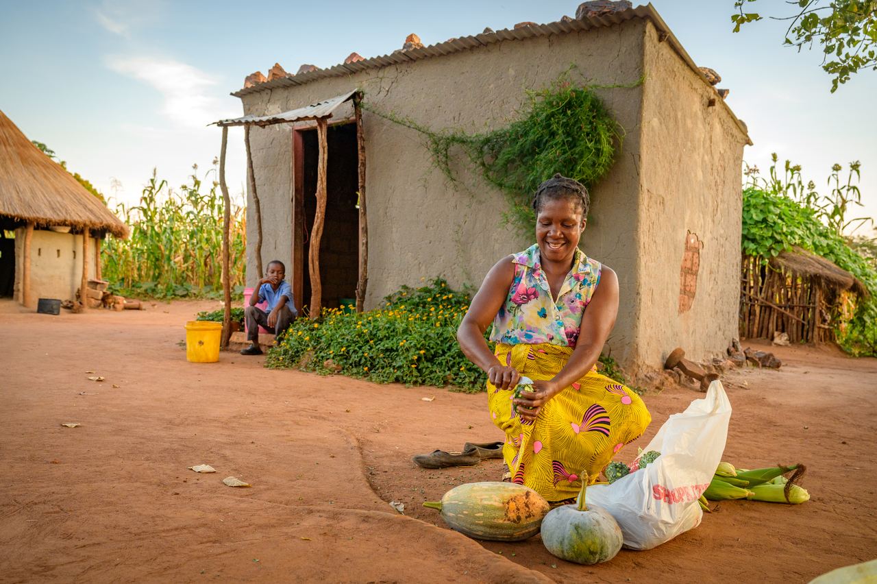 A farmer kneels in the yard of a homestead sorting pumpkins, maize and a bag of produce, with another person seated behind.